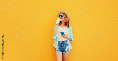 Happy smiling young woman 20s drinking coffee with smartphone looking away wearing summer straw hat, shorts on orange background