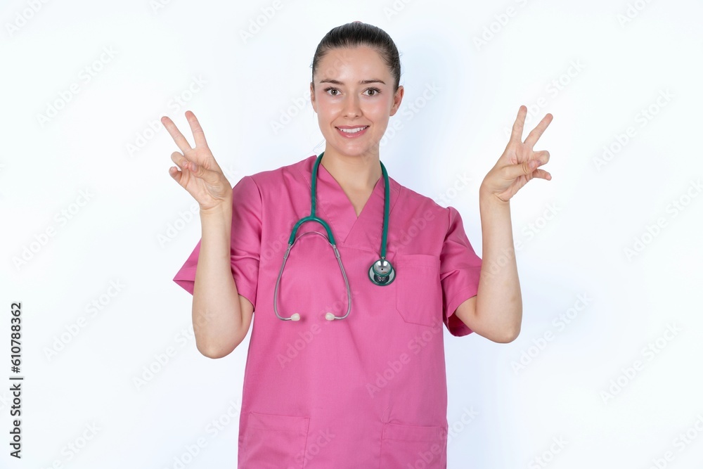 young caucasian doctor woman wearing pink uniform over white background with optimistic smile, showing peace or victory gesture with both hands, looking friendly. V sign. young caucasian doctor woman wearing pink uniform over white background with optimistic smile, showing peace or victory gesture with both hands, looking friendly. V sign.