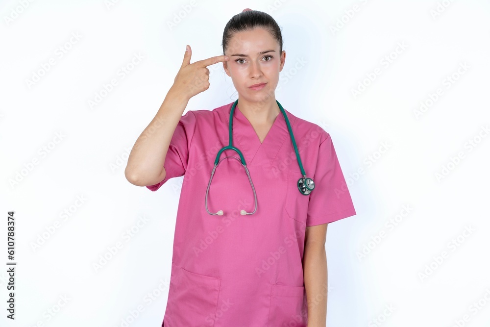 Unhappy young caucasian doctor woman wearing pink uniform over white background curves lips and makes suicide gesture, shoots in temple with hand, tries kill himself. Unhappy young caucasian doctor woman wearing pink uniform over white background curves lips and makes suicide gesture, shoots in temple with hand, tries kill himself.