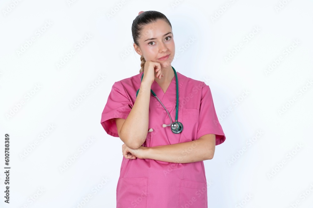 Face expressions and emotions. Thoughtful young caucasian doctor woman wearing pink uniform over white background holding hand under his head, having doubtful look. Face expressions and emotions. Thoughtful young caucasian doctor woman wearing pink uniform over white background holding hand under his head, having doubtful look.