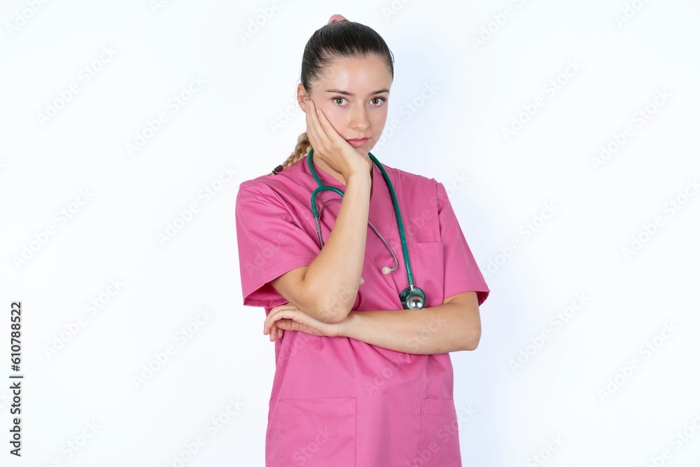 Very bored young caucasian doctor woman wearing pink uniform over white background holding hand on cheek while support it with another crossed hand, looking tired and sick. Very bored young caucasian doctor woman wearing pink uniform over white background holding hand on cheek while support it with another crossed hand, looking tired and sick.