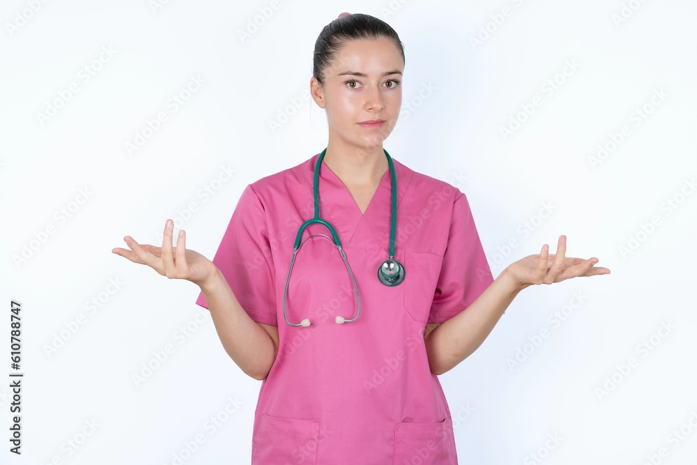 young caucasian doctor woman wearing pink uniform over white background looks uncertain shrugs shoulders. young caucasian doctor woman wearing pink uniform over white background looks uncertain shrugs shoulders.