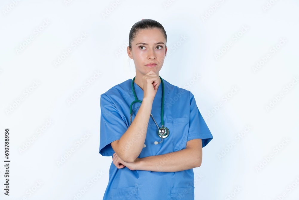 Thoughtful young caucasian doctor woman wearing medical uniform over white background holds chin and looks away pensively makes up great plan