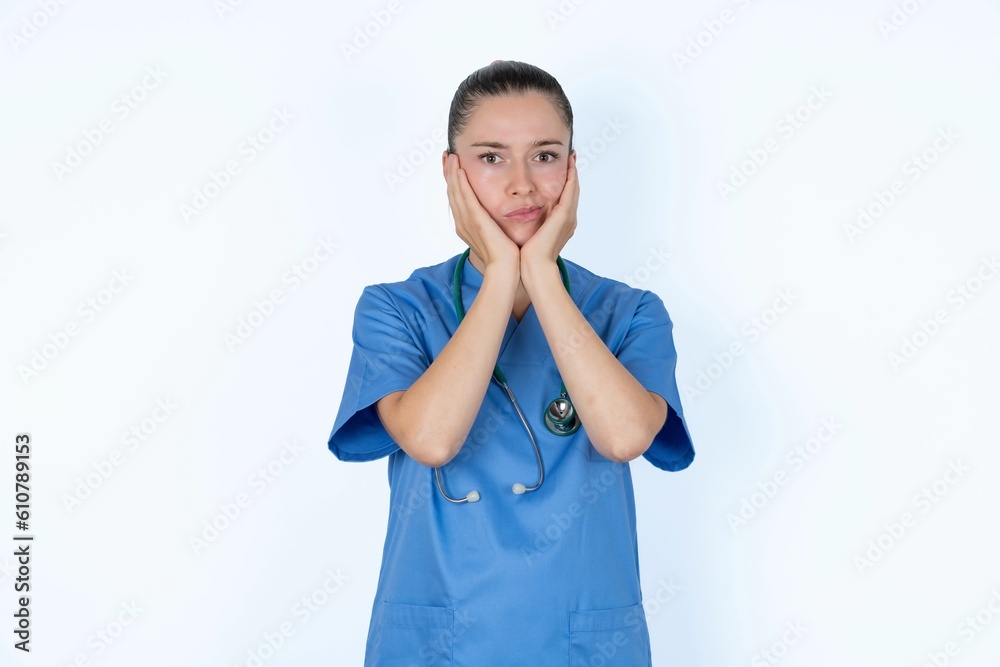 young caucasian doctor woman wearing medical uniform over white background with surprised expression keeps hands under chin keeps lips folded makes funny grimace young caucasian doctor woman wearing medical uniform over white background with surprised expression keeps hands under chin keeps lips folded makes funny grimace