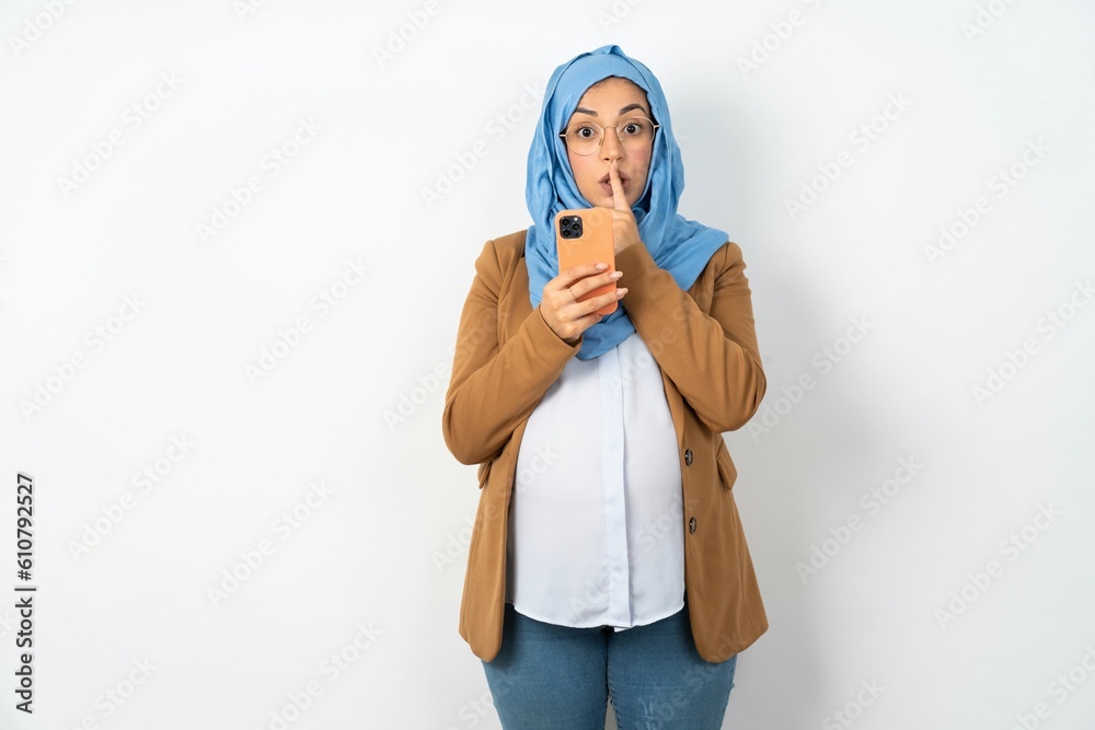 Young beautiful pregnant muslim woman wearing hijab over white background holding modern gadget ask not tell secrets