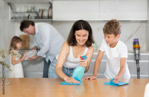 Positive woman washes kitchen table by rag with her son. Husband and daughter help to do cleaning in kitchen in background. Joint family house cleaning, household tasks, household help