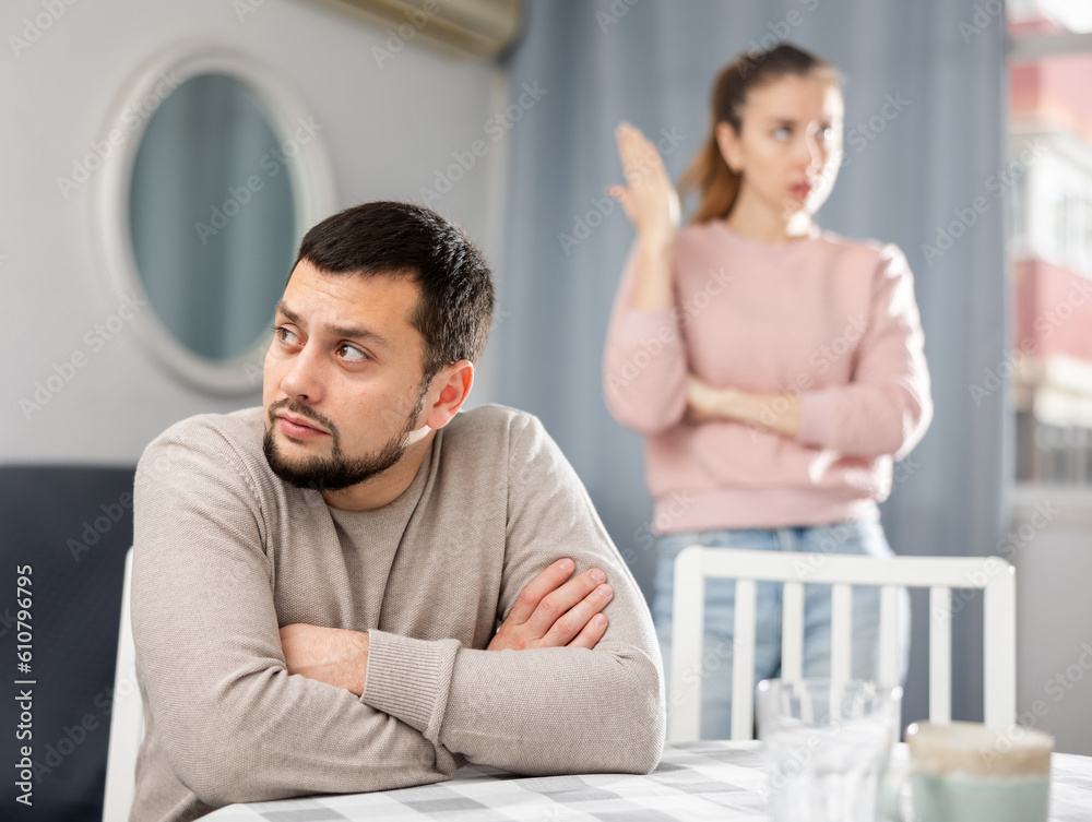 Obraz premium Sad man sitting at table in apartment while his wife standing behind and quarreling with him.