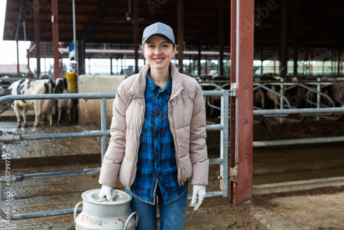 Portrait of young adult smiling woman farm worker holding big milk can at cowshed on dairy farm