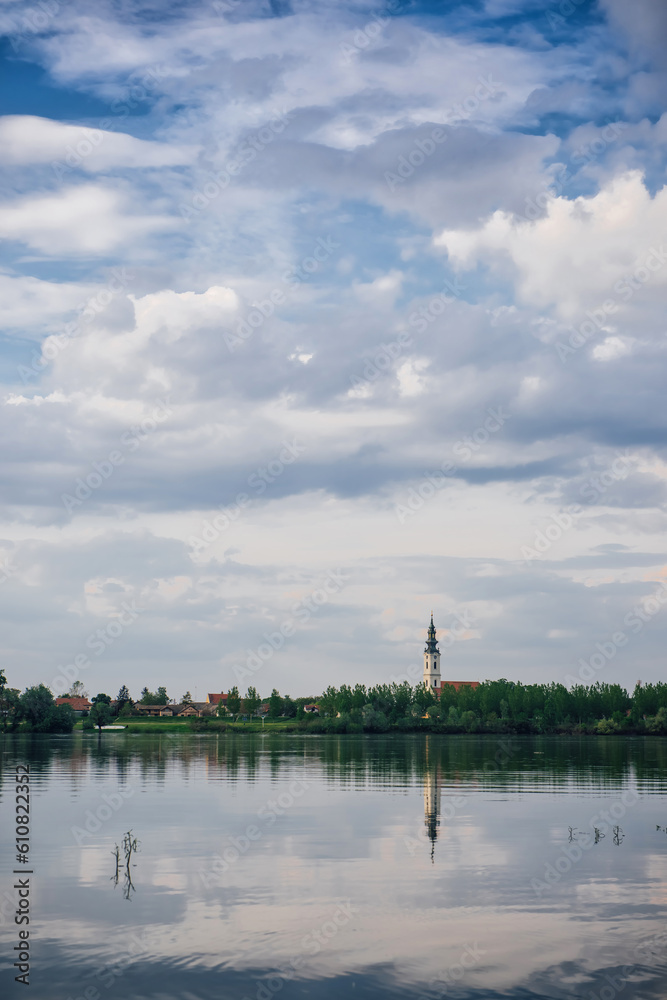 Rural view of the old cathedral tower with reflection in the water and cottages against the background of a wide river and cloudy sky. Balkan view of the Danube Rural view of the old cathedral tower with reflection in the water and cottages against the background of a wide river and cloudy sky. Balkan view of the Danube
