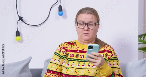 Dissatisfied bored woman grouch in glasses, ugly sweater sits at party on couch holds smartphone in her hands, scrolls tape, looks at people around, rolls her eyes dismissively. Family dinner phubbing photo