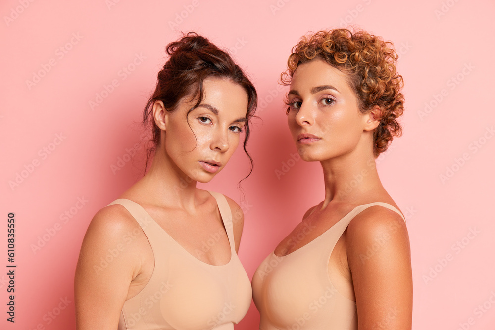 Photo portrait of two beautiful women half turned to each other looking at the camera with serious faces dressed in beige sports bras, friendship oncept, copy space