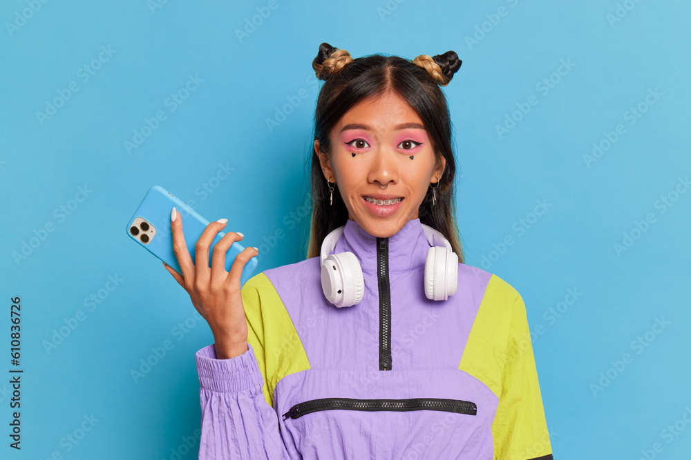 Oriental smiling girl with funny make-up wearing violet-colored sweatshirt having headphones on her neck, holds phone in hand, stands on blue background, copy space