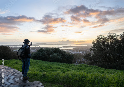 Tourist taking photo using smartphone at Mt Eden summit at sunrise. Rangitoto Island in the distance. Auckland.