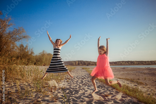 Happy mother and daughter in dresses are jumping and dancing on the beach during sunset. A good relation between generations. Fun family pastime. Health promotion through outdoor activities. Vacation