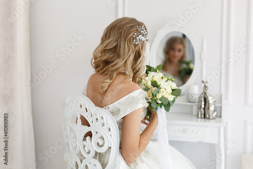 Beautiful young bride sitting on chair and looks at herself in mirror in beauty salon with white interior. blonde woman in white dress with hair style and makeup  looking in mirror back view portrait