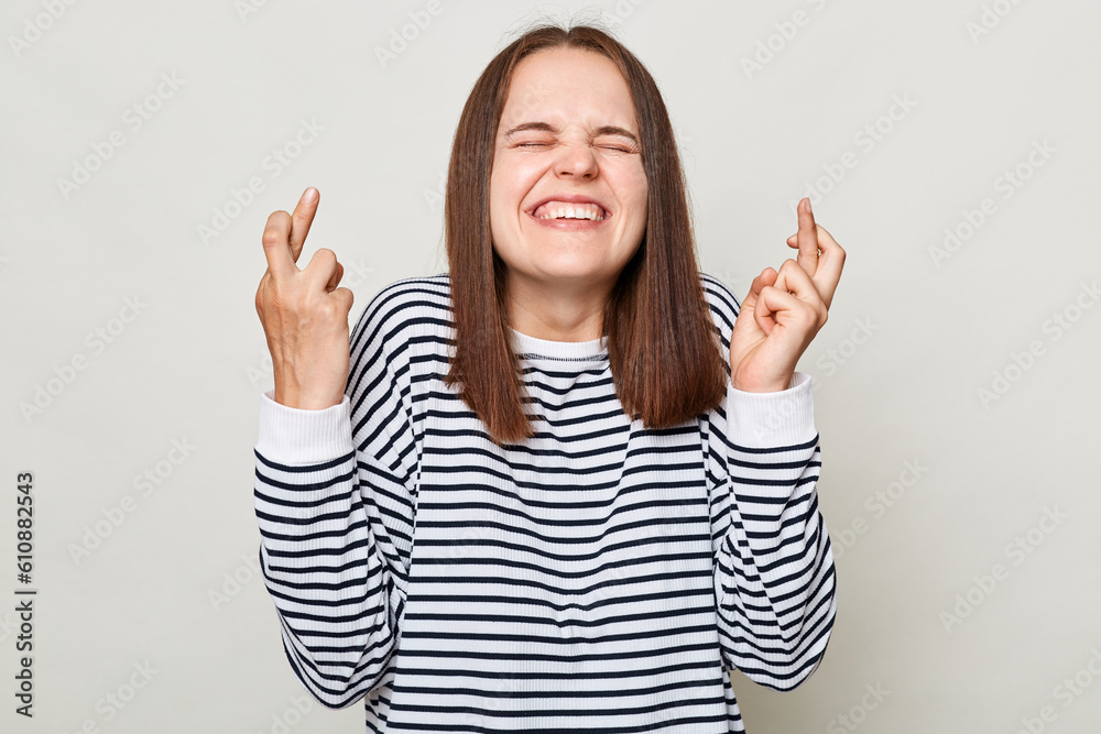 Making wish. Praying for good luck. Smiling hopeful brown haired woman wearing striped shirt posing isolated over gray background standing with crossed fingers and closed eyes. Making wish. Praying for good luck. Smiling hopeful brown haired woman wearing striped shirt posing isolated over gray background standing with crossed fingers and closed eyes.