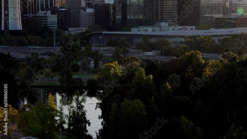 Dawn  over Perth  City with speeding traffic and crystal clear pond surrounded by trees in relatively dark foreground. Golden glow of leaves as sunlight emerges