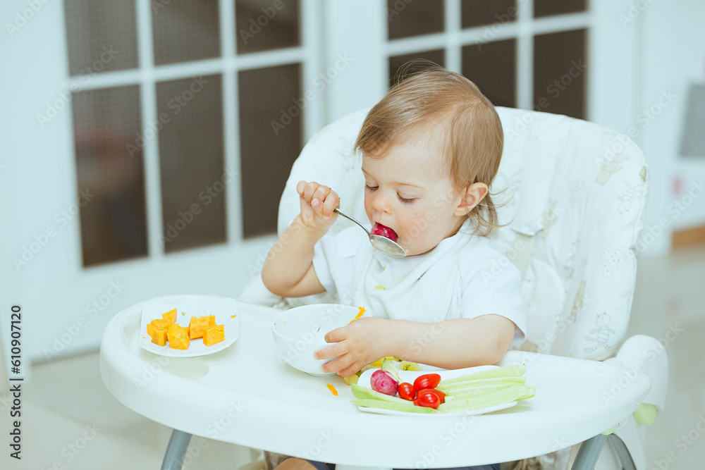 Little toddler eating using spoon in hand, learning how to feed himself. Infant sitting on child chair, eating from white bowl, trying healthy food lunch time at home baby led weaning Little toddler eating using spoon in hand, learning how to feed himself. Infant sitting on child chair, eating from white bowl, trying healthy food lunch time at home baby led weaning