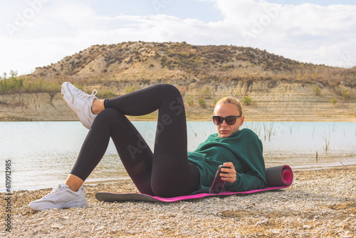 a girl of European appearance in sportswear lies on a yoga mat near a beautiful lake. Beautiful landscape of nature, there is a place for an inscription. Lake with turquoise water.