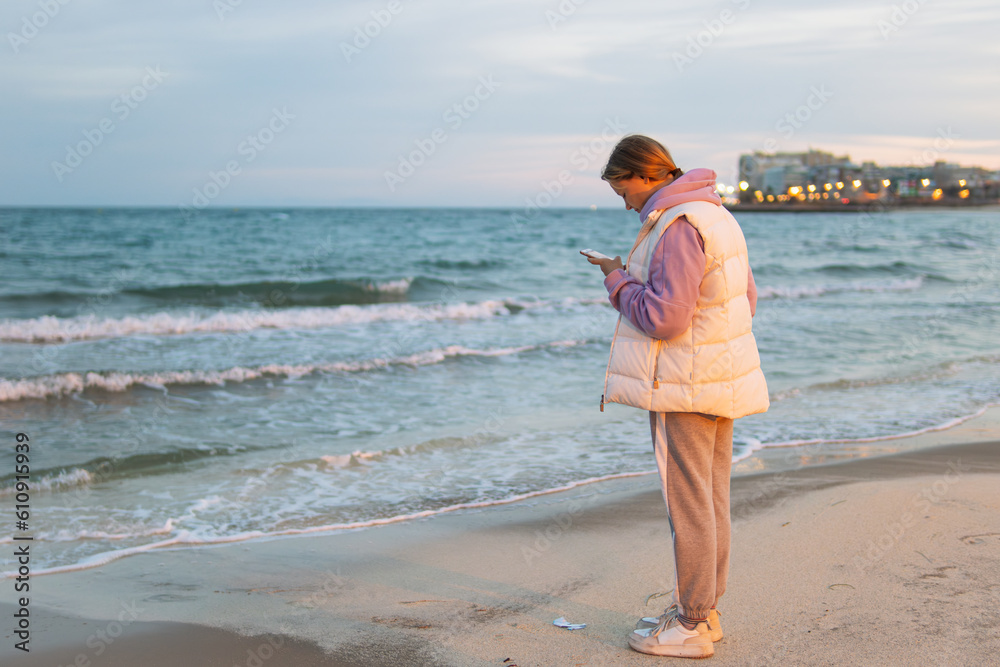 A girl of European make in casual clothes stands near the ocean with a phone in her hands is sad and looks into the phone.