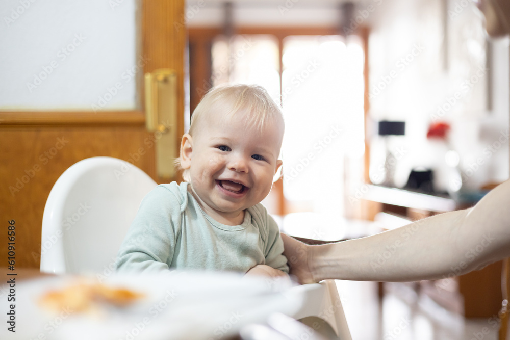 Adorable cheerful happy infant baby boy child smiling while sitting in high chair at the dining table in kitchen at home beeing spoon fed by his mother. Adorable cheerful happy infant baby boy child smiling while sitting in high chair at the dining table in kitchen at home beeing spoon fed by his mother.