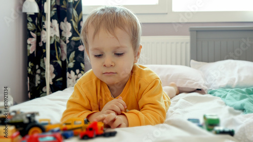 Little baby boy lying in bed and driving toy cars over the pillow. Child development, education and entertainment
