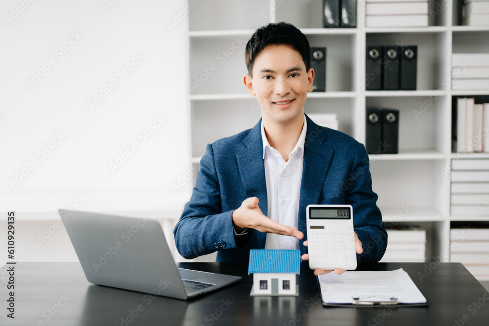 Young real estate agent worker working with laptop and tablet at table in modern office and small house beside it.. Young real estate agent worker working with laptop and tablet at table in modern office and small house beside it..