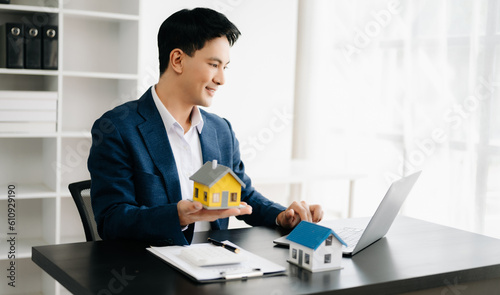 Young real estate agent worker working with laptop and tablet at table in modern office and small house beside it..