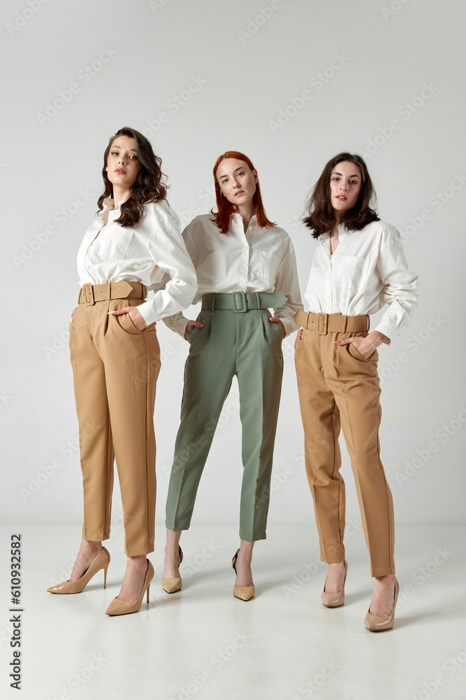 Full-length portrait of three beautiful women in stylish clothes, pants and blouses standing against light studio background. Concept of elegant fashion, female beauty, business, shopping, sales, ad Full-length portrait of three beautiful women in stylish clothes, pants and blouses standing against light studio background. Concept of elegant fashion, female beauty, business, shopping, sales, ad