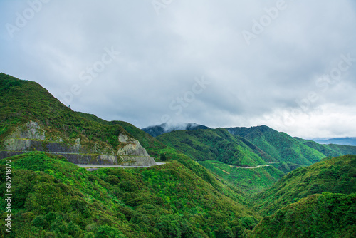 Mountain road winding along a green gorge with forest in the mountains. Aerial view over green hills, trees, road, and steep cliffs. Low clouds over Remutaka Crossing, North Island, New Zealand