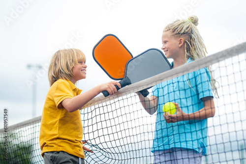 Laughing boy and girl playing pickleball game, hitting pickleball yellow ball with paddle, outdoor sport leisure kids activity.