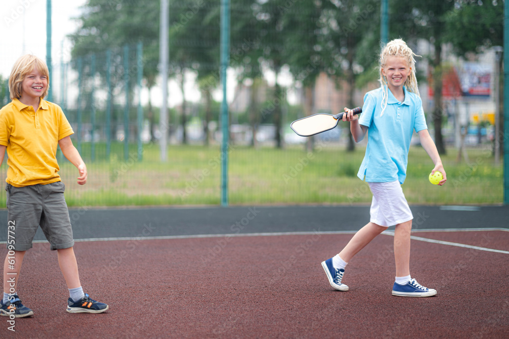 Laughing boy and girl playing pickleball game, hitting pickleball yellow ball with paddle, outdoor sport leisure kids activity.