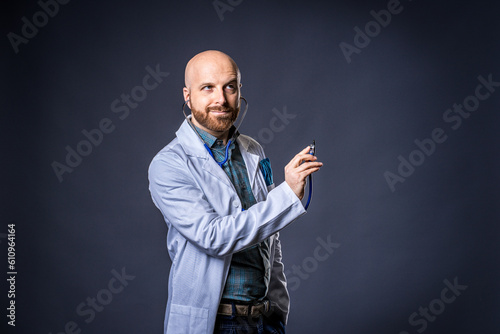 Photo of doctor with beard and stethoscope posing listening with blue background and medical white coat