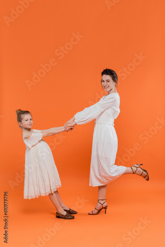 summer trends, young mother holding hands with preteen daughter and standing on orange background, white sun dresses, togetherness, fashion and style concept, bonding, joyful