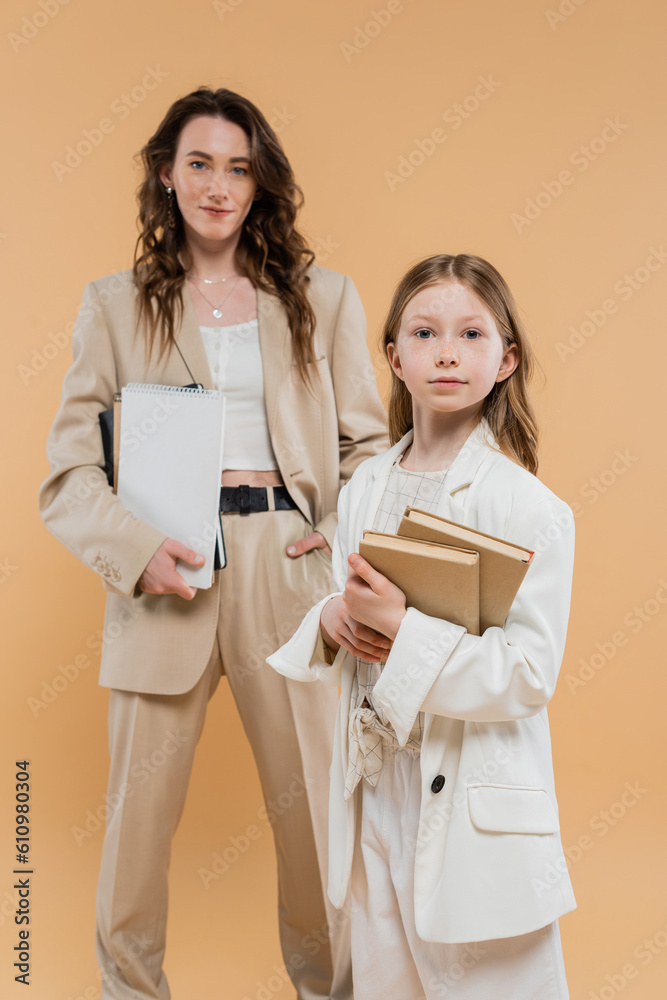Fototapeta premium trendy mother and daughter in suits, child holding books near blurred woman with notebooks on beige background, fashionable outfits, formal attire, corporate mom, education concept
