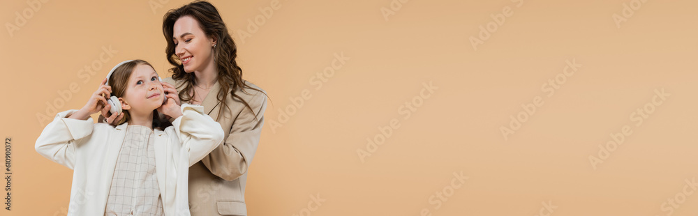 corporate mom and daughter in suits, happy woman wearing wireless headphones on girl while standing together on beige background, fashionable outfits, formal attire, modern family, banner corporate mom and daughter in suits, happy woman wearing wireless headphones on girl while standing together on beige background, fashionable outfits, formal attire, modern family, banner