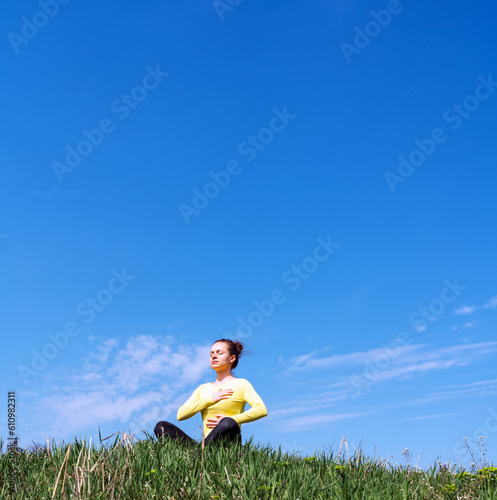 Yoga breathing exercise outdoors in nature, woman meditating on green grass against blue sky.
