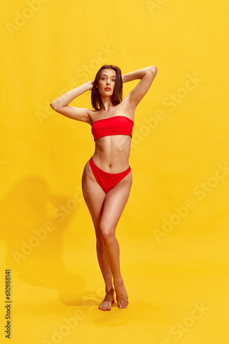 Attractive, brunette young girl with relief, fit, perfect body, in red swimsuit posing against yellow studio background © Lustre Art Group 