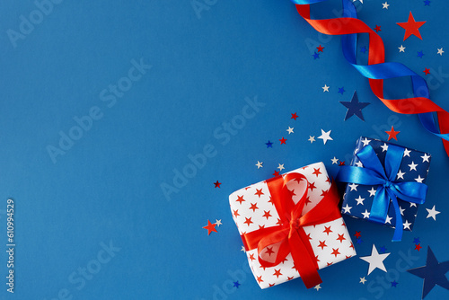 American Independence Day concept. Top view flat lay of patriotic gift boxes, ribbons and festive confetti on blue background with empty space for text or greeting
