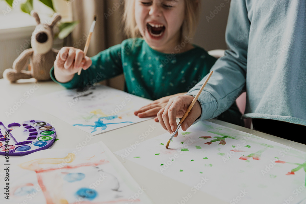 Two cute little sisters or classmates are painting on table with brush. Small girls are laughing. Kids are in kindergarten or at home. Child in living room. Home schooling concept, doing homework 