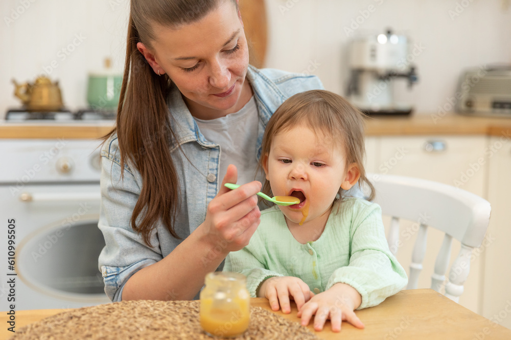 Happy family at home. Mother feeding her baby girl from spoon in kitchen. Little toddler child with messy funny face eats healthy food at home. Young woman mom giving food to kid daughter Happy family at home. Mother feeding her baby girl from spoon in kitchen. Little toddler child with messy funny face eats healthy food at home. Young woman mom giving food to kid daughter