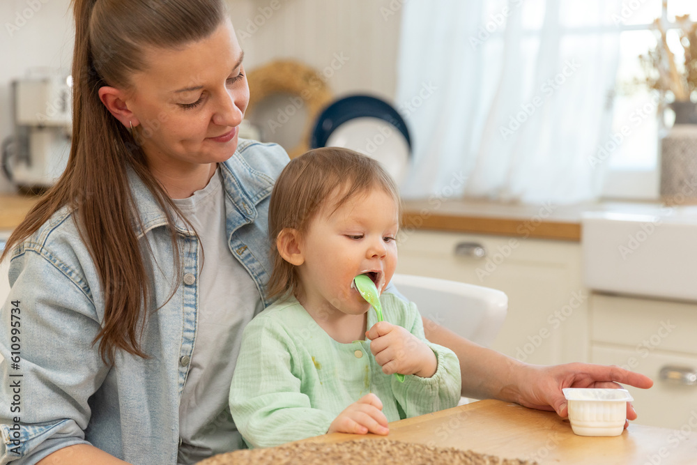 Happy family at home. Mother feeding her baby girl from spoon in kitchen. Little toddler child with messy funny face eats healthy food at home. Young woman mom giving food to kid daughter Happy family at home. Mother feeding her baby girl from spoon in kitchen. Little toddler child with messy funny face eats healthy food at home. Young woman mom giving food to kid daughter