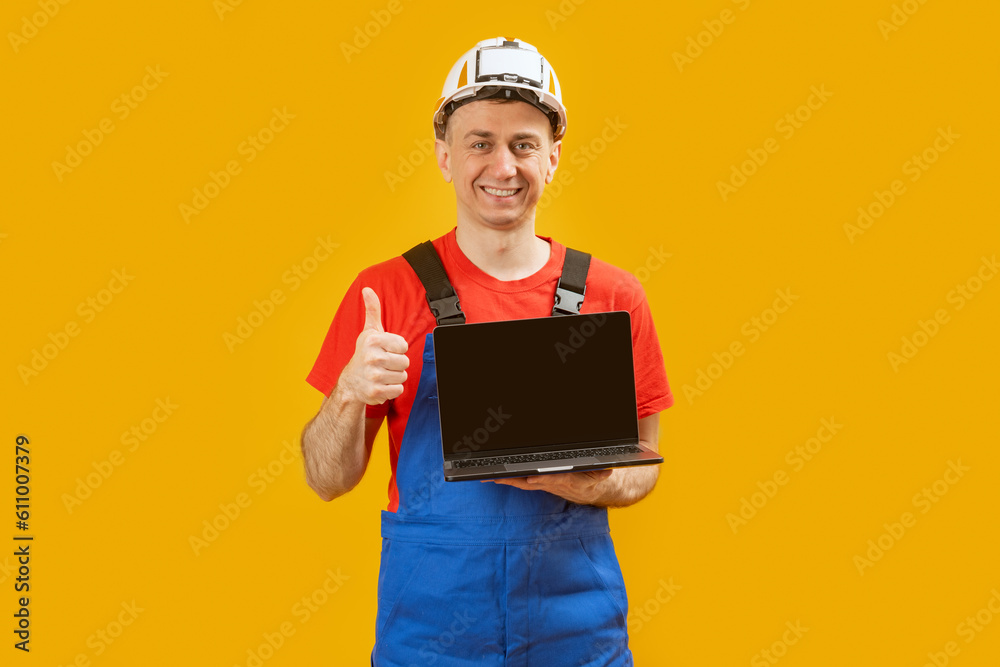 Man in blue jumpsuit and safety helmet holds laptop with blank black screen and shows thumbs up, gesture like. Copy space, mock up. Man in blue jumpsuit and safety helmet holds laptop with blank black screen and shows thumbs up, gesture like. Copy space, mock up.