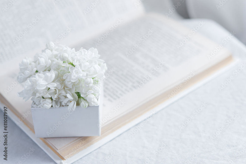 Close-up of a bouquet of white flowers in a white gift box on an open book on a white textile background. Slow life concept. Greeting card mockup	 - obrazy, fototapety, plakaty 