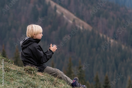 Blond boy in mountains sitting on hillside and drinking tea from cup. Hiking with child, resting in mountains. Active recreation.