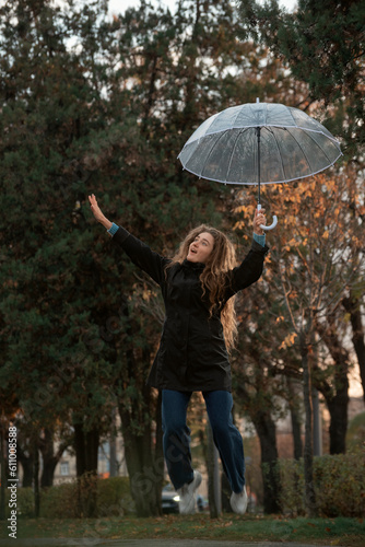 Pretty young woman is photographed jumping with transparent umbrella in hand. Girl with long curly hair in autumn park