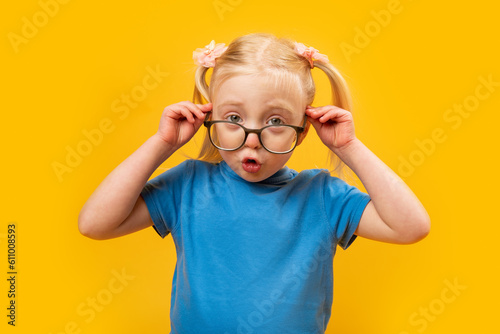Surprised girl of 5-6 years old adjusts her glasses. Portrait of small blonde girl in blue T-shirt with glasses on yellow background.