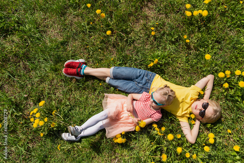 Little boy and his younger sister are lying on green field with dandelions. Siblings rest in the grass. Top view
