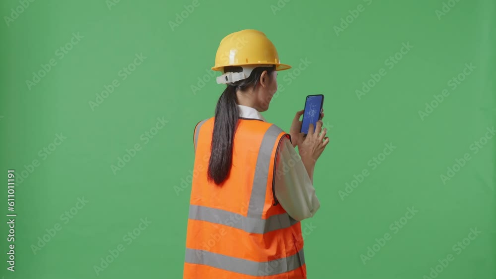 Back View Of Asian Female Engineer With Safety Helmet Looking At The Wind Turbine Blueprint On Smartphone While Standing In The Green Screen Background Studio
