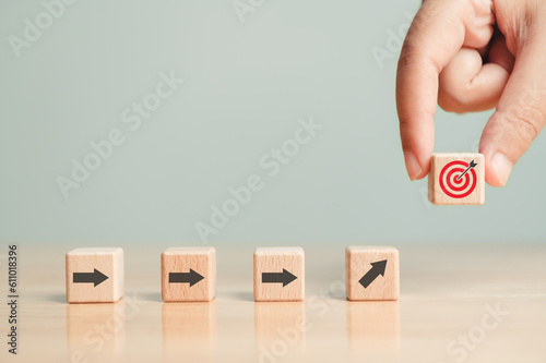 Obraz na płótnie Arrow leading point to target icon on wooden blocks with businessman hand placing red goal dartboard symbol